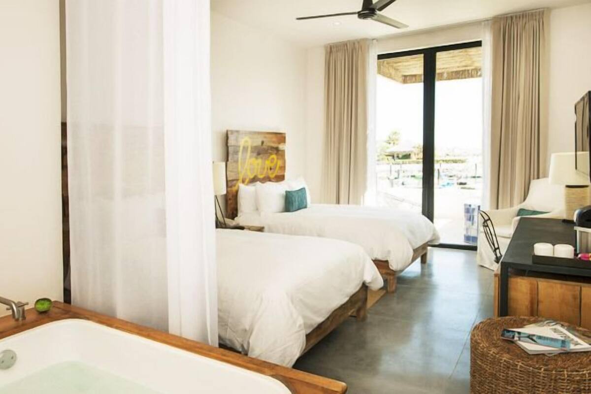 A hotel room with two beds, a desk with a lamp, a window with curtains, and a view of the outdoors.