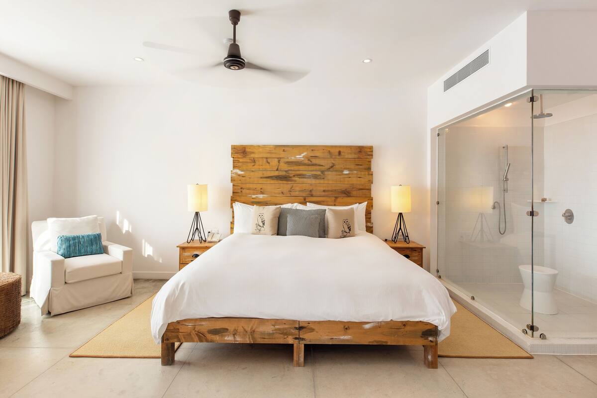 A bedroom with a wooden headboard, a bed with white bedding, two bedside lamps, a wooden nightstand, a chair, and a glass-enclosed shower area.