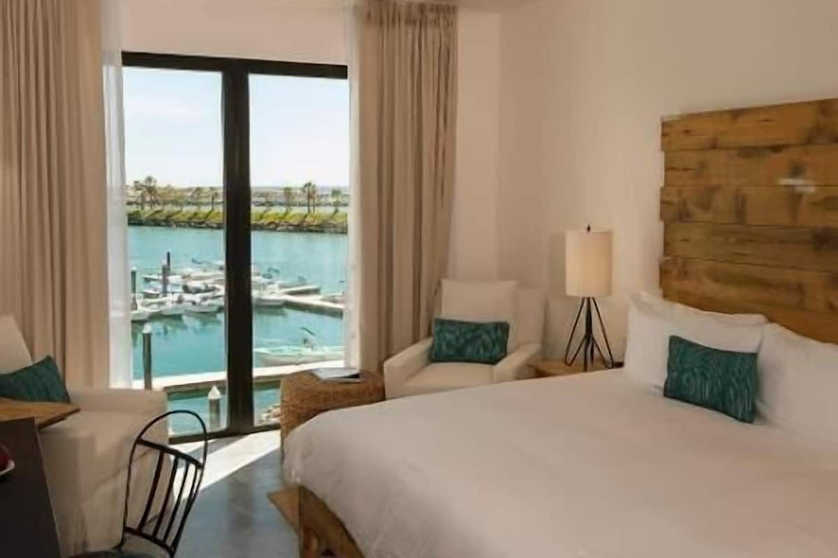 A hotel room with a bed, a desk, a chair, a lamp, and a view of the marina.