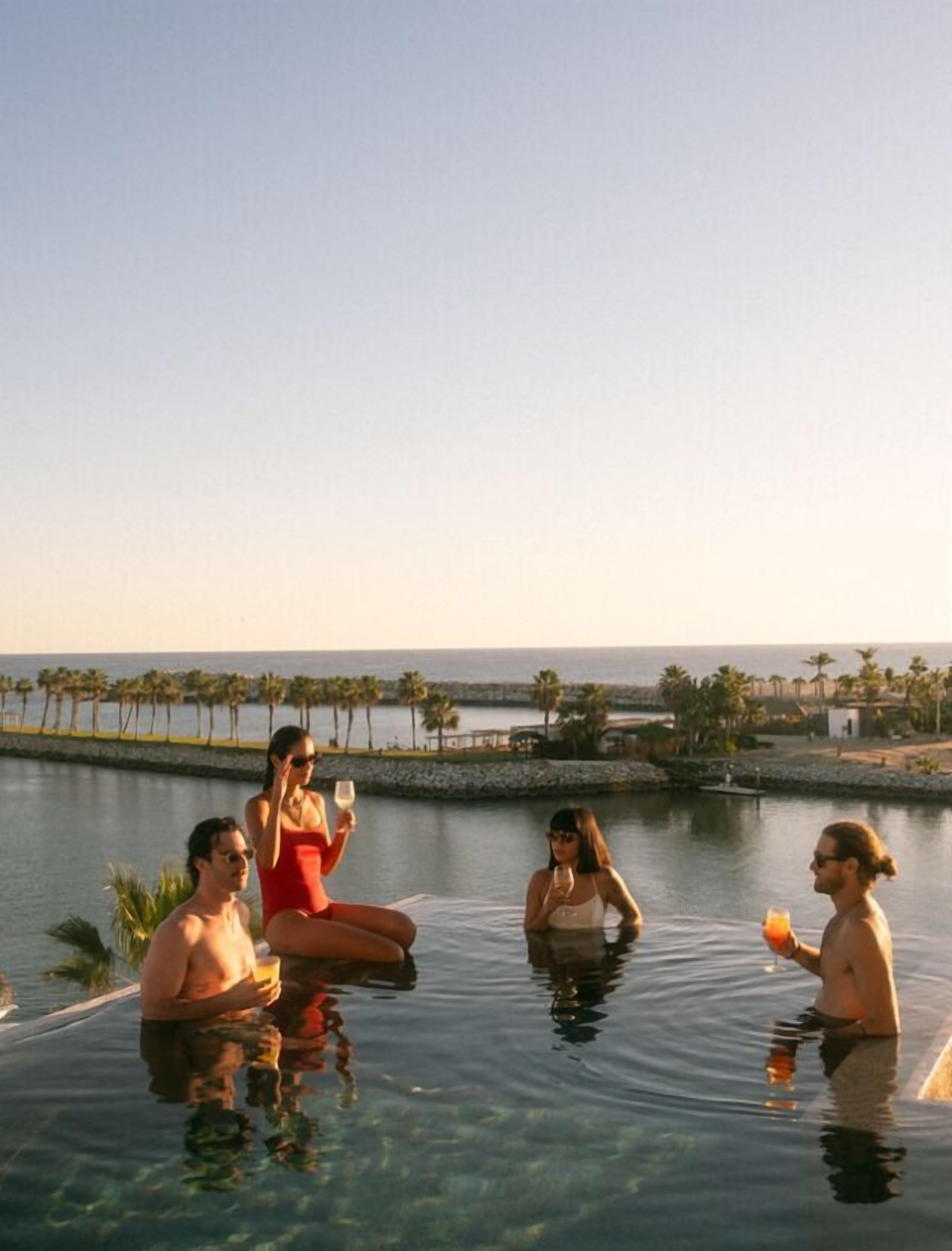 Um grupo de pessoas relaxando em uma piscina cristalina com vista para o oceano e palmeiras.