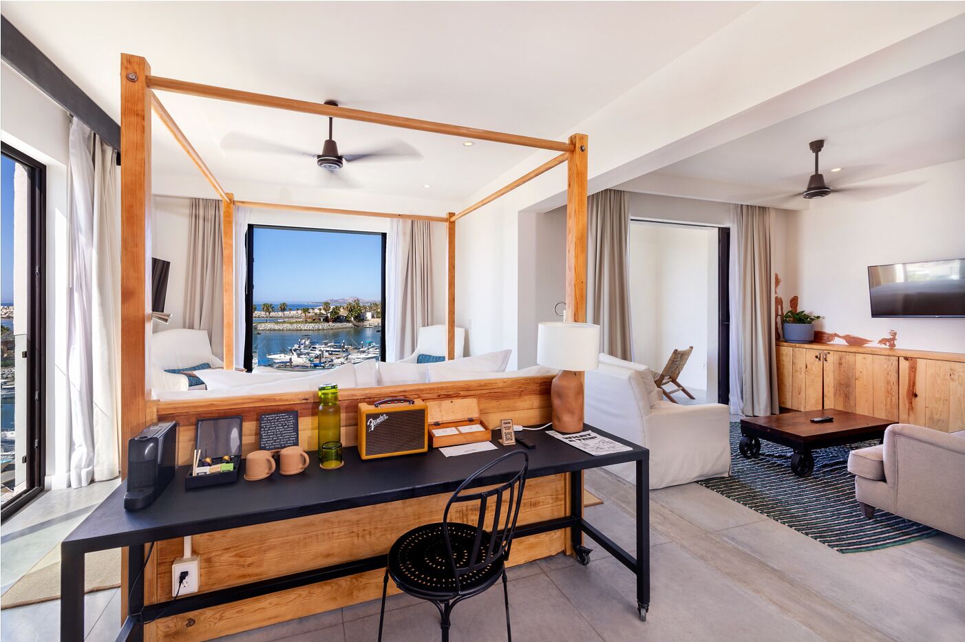 A modern hotel room with a large window overlooking a marina, a wooden desk with a chair, a sofa, and a television on the wall.