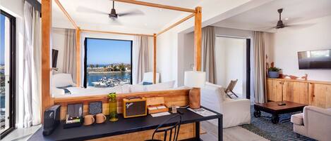 A modern hotel room with a large window overlooking a marina, a wooden desk with a chair, a sofa, and a television on the wall.