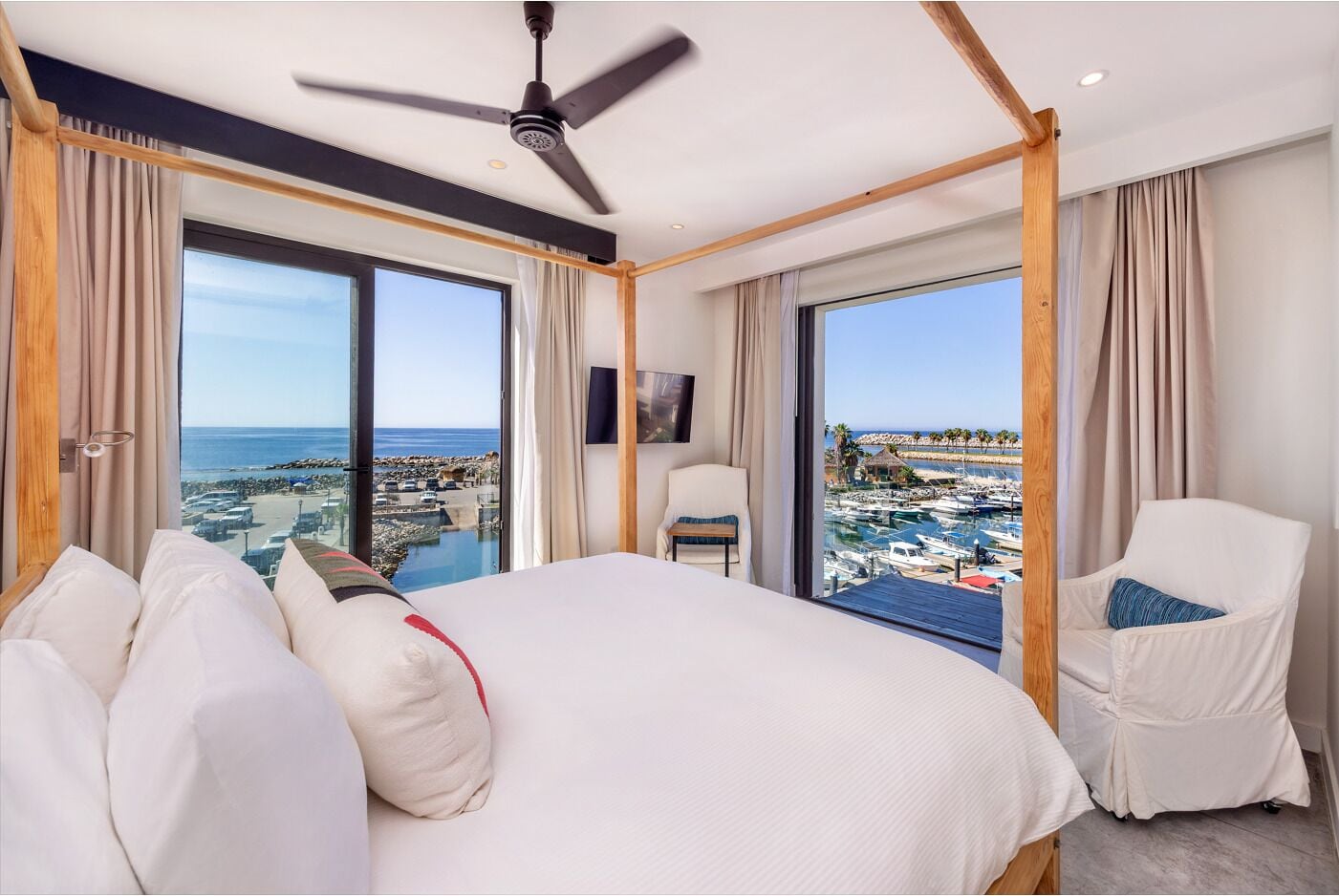 Une chambre d’hôtel avec un grand lit, un ventilateur de plafond, une chaise et une vue sur un port de plaisance et une plage.