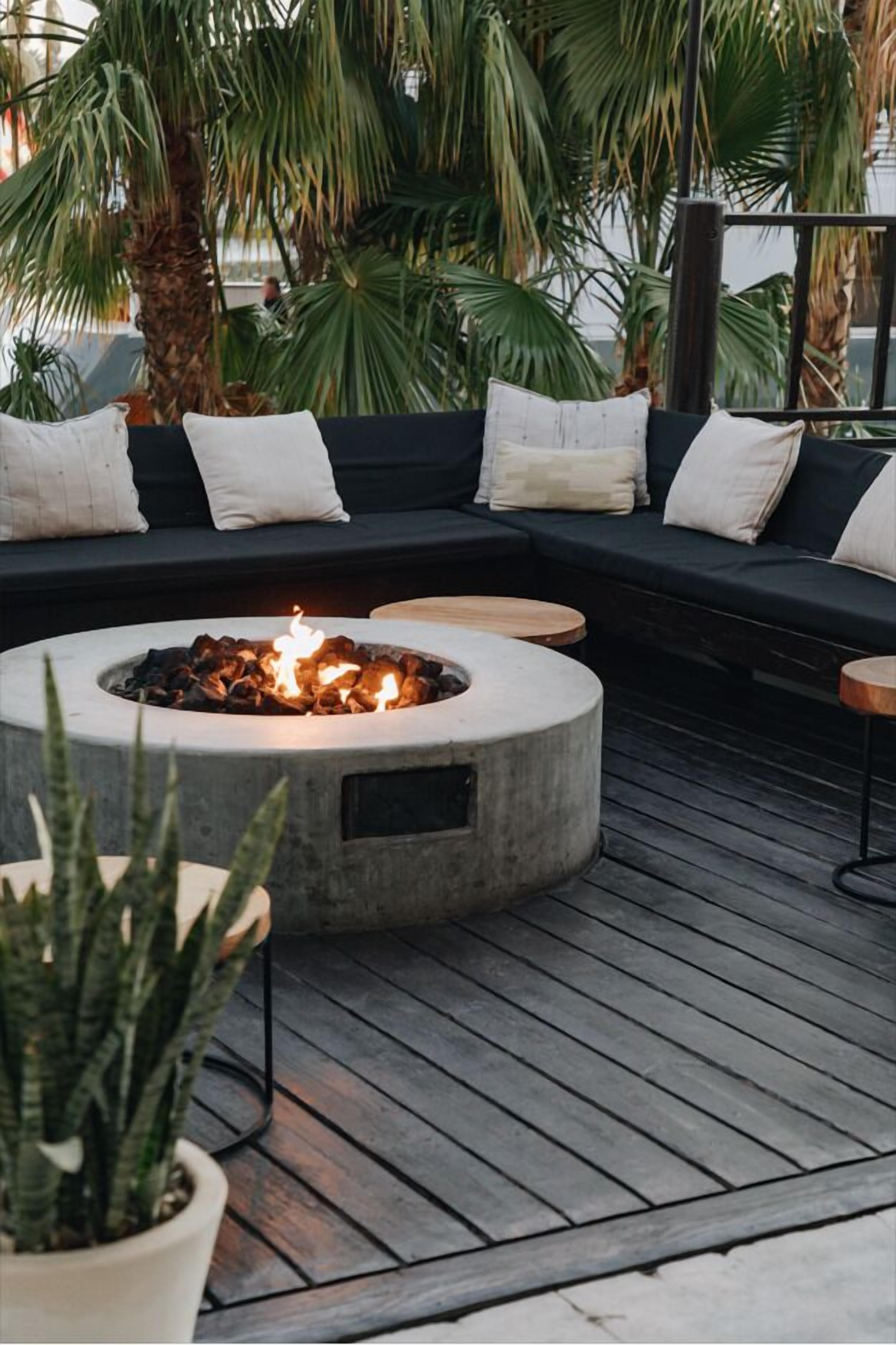 A cozy outdoor seating area with a circular fire pit, a dark-colored sofa with cushions, and potted plants.