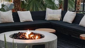 A cozy outdoor seating area with a circular fire pit, a dark-colored sofa with cushions, and potted plants.