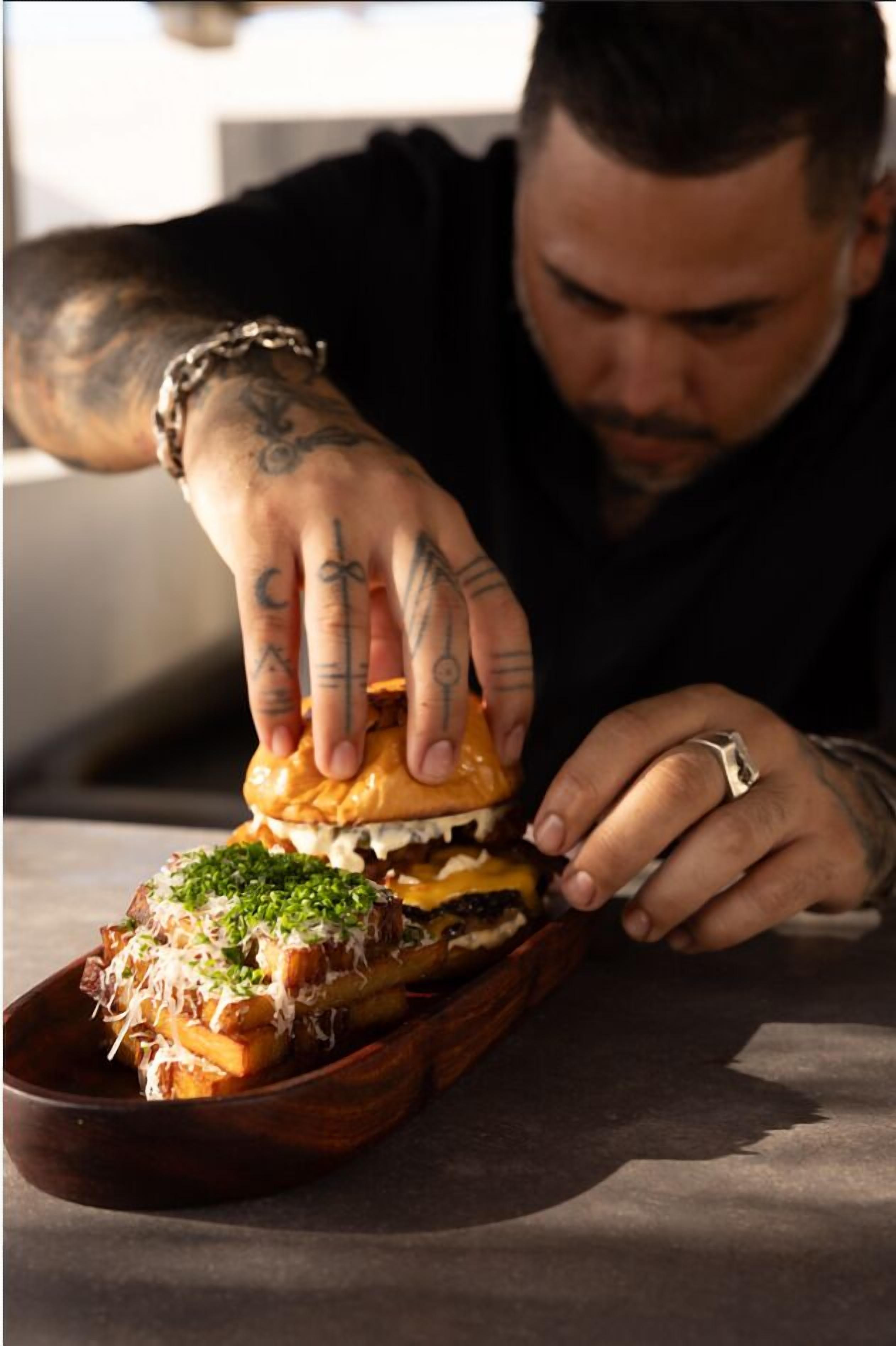 A person with tattoos on their hands holding a sandwich with a bun, cheese, and green garnish.