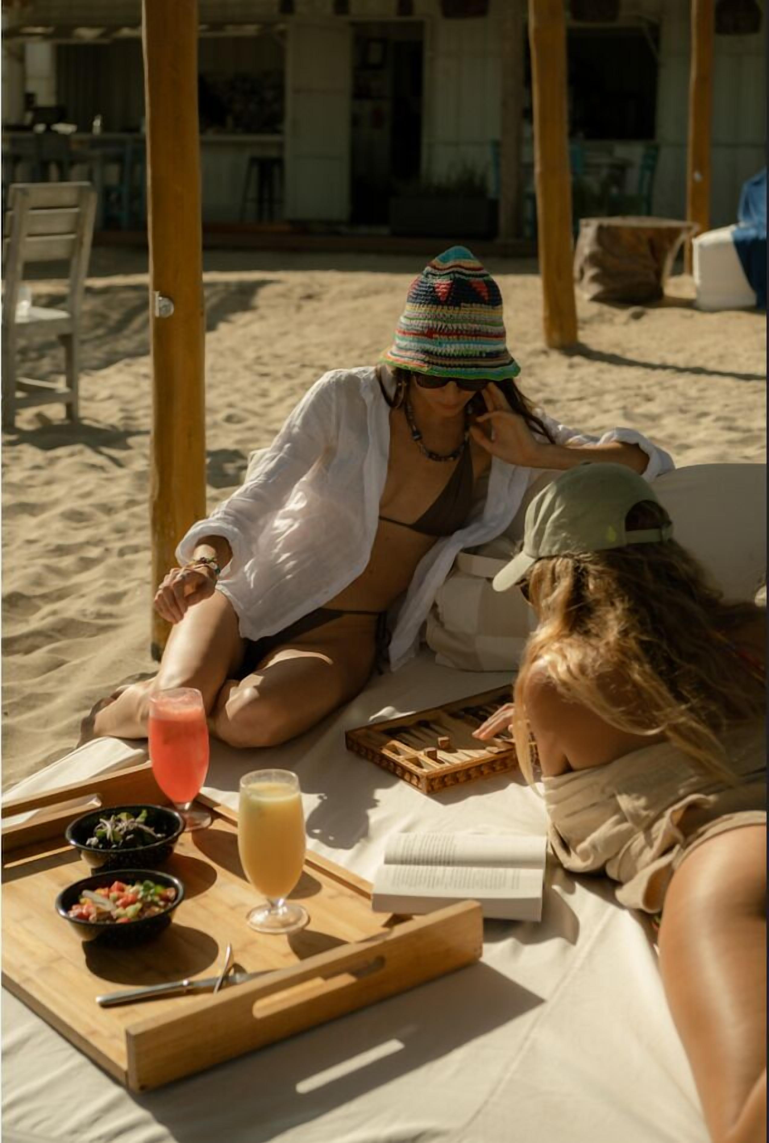 Una spiaggia con un tavolo di legno apparecchiato per un pasto, bevande e un libro.