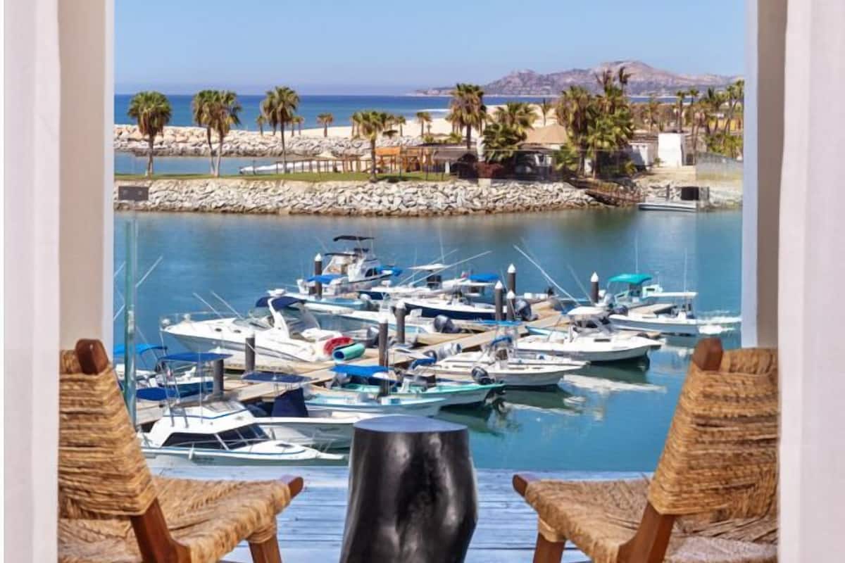 A balcony with two wicker chairs, a black table, and a view of a marina with boats and palm trees.
