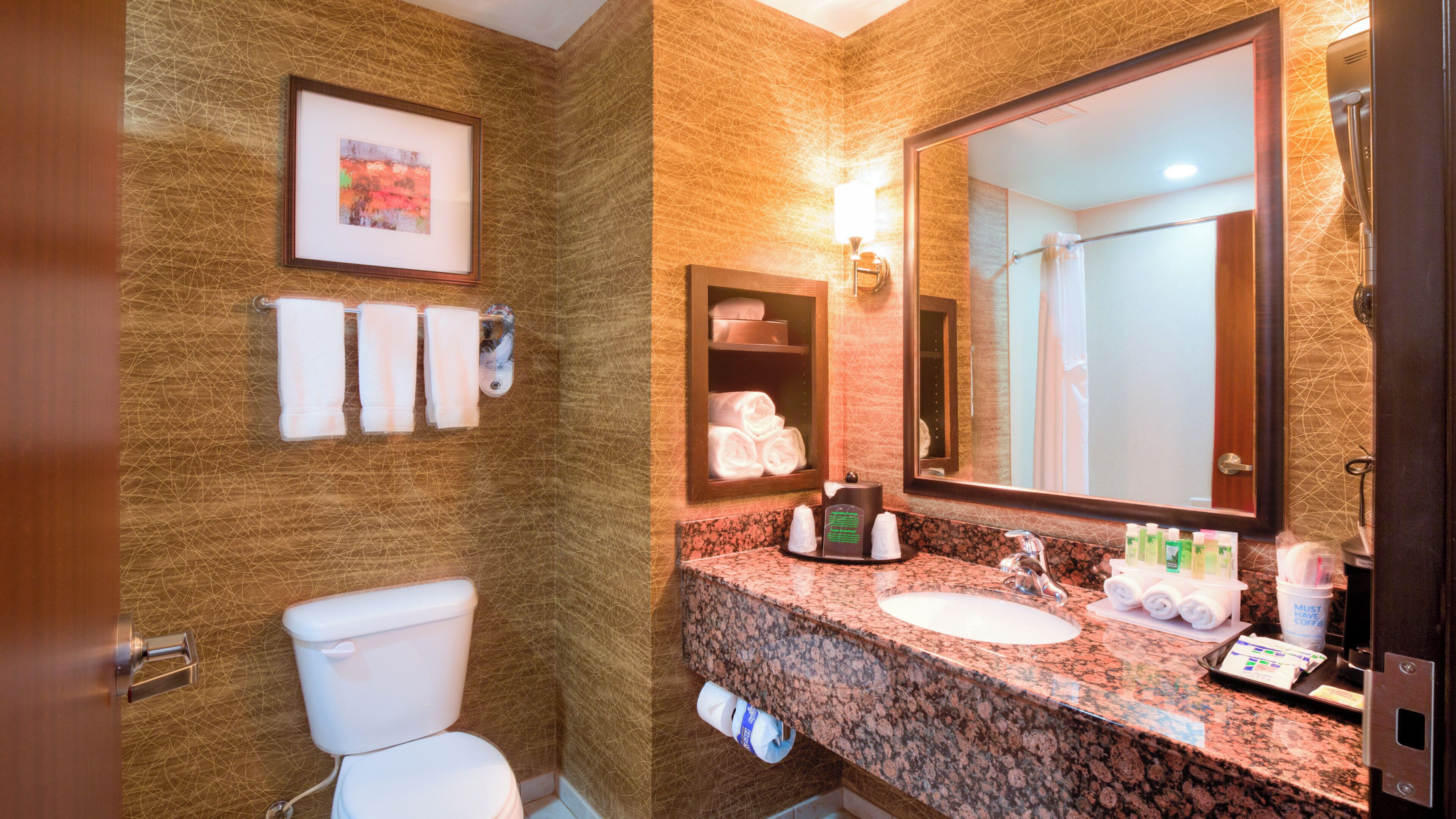 Une salle de bain avec des toilettes, un lavabo et un miroir. Des serviettes sont accrochées au mur et un tableau encadré est suspendu au-dessus des toilettes.