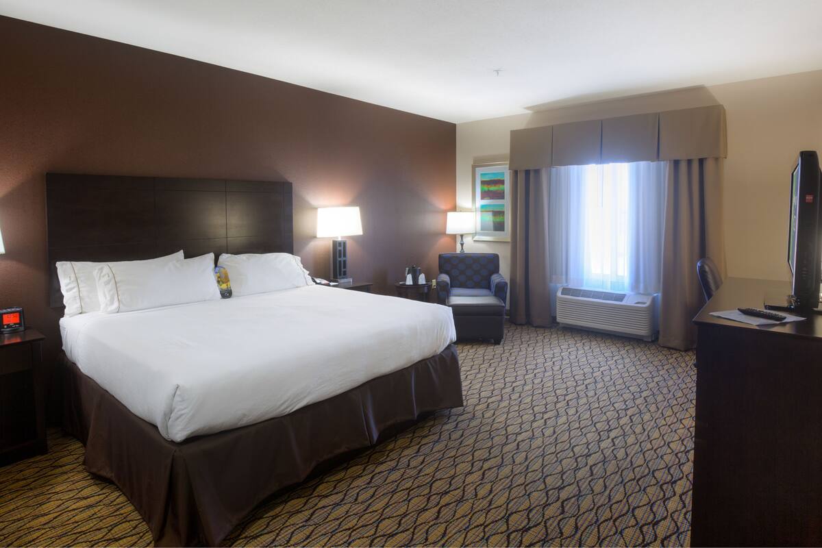 Ein Hotelzimmer mit einem großen Bett, zwei Nachttischen, einem Schreibtisch mit Computer, einem Sessel und einem Fenster mit Vorhängen.