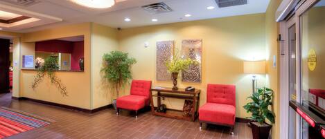 A hotel lobby with a reception desk, two red chairs, a potted plant, and a wall with abstract art.