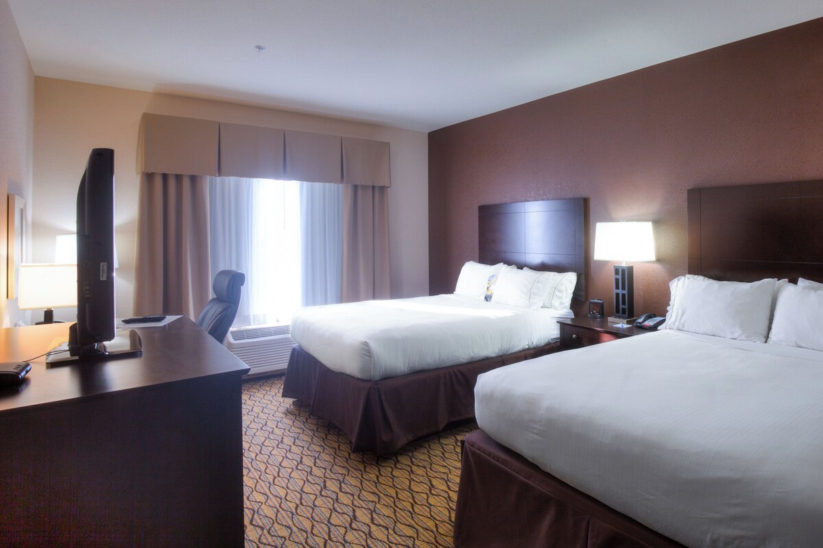 Ein Hotelzimmer mit zwei Betten, einem Schreibtisch mit Computer, einem Sessel, einem Fernseher und einem Fenster mit Vorhängen.