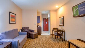 A hotel room with a striped sofa, a polka dot armchair, a small table, and a bench. There is a kitchenette area with a coffee machine and a refrigerator. A door leads to a hallway with a framed picture on the wall. - Holiday Inn Express Hotel & Suites Elk City by IHG (Elk City)