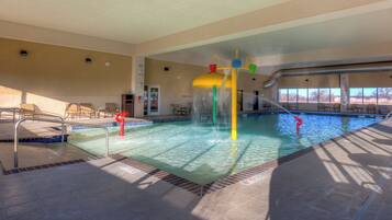 An indoor swimming pool with multiple water features, including a central fountain, surrounded by lounge chairs and a tiled deck.