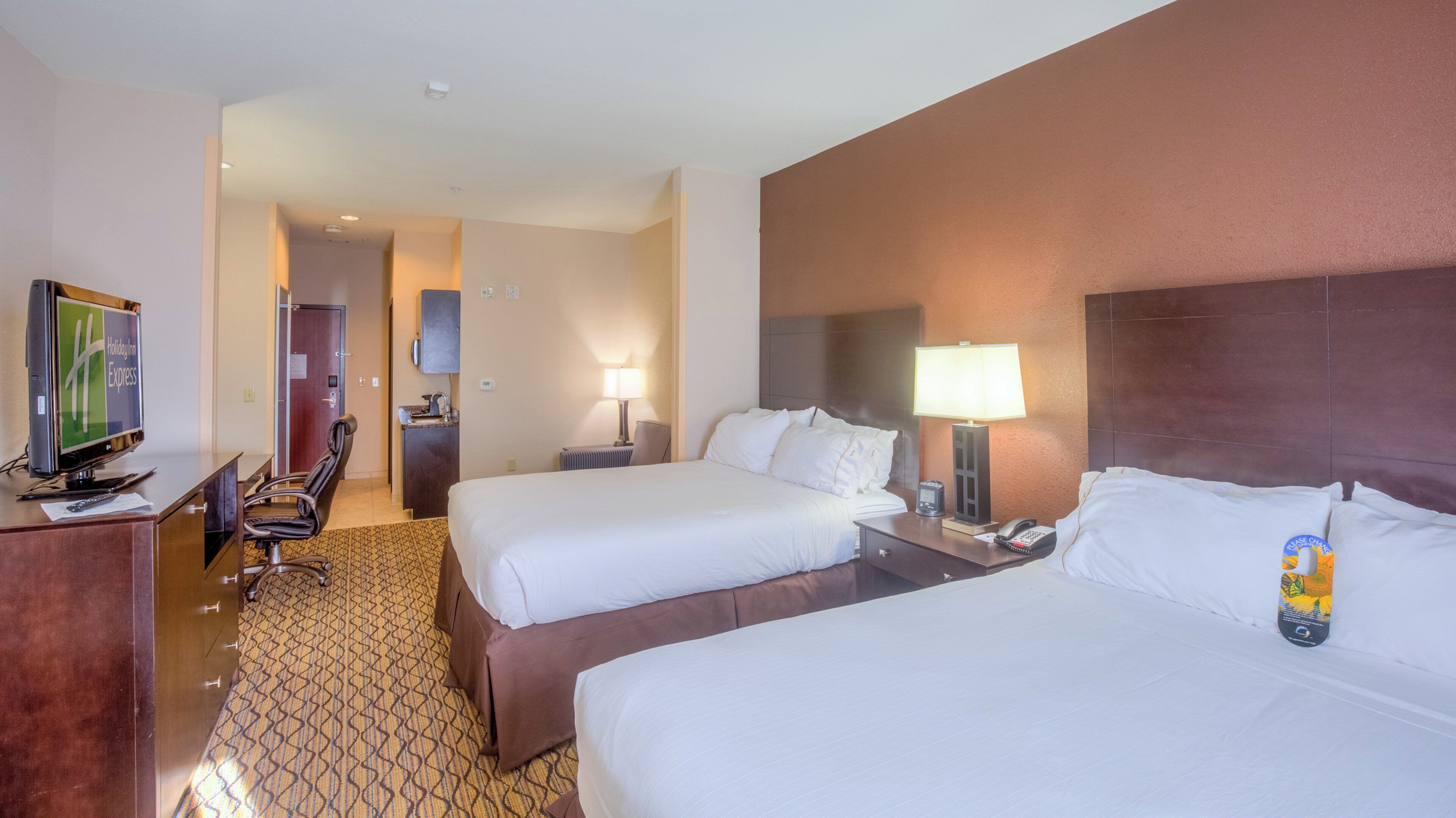 A hotel room with two beds, a desk with a TV, a chair, a desk lamp, and a bedside table with a phone and a notepad.