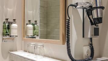 Une salle de bain équipée d’un sèche-cheveux fixé au mur, d’un miroir et d’un plan de toilette en marbre agrémenté de pots et de bouteilles en verre.