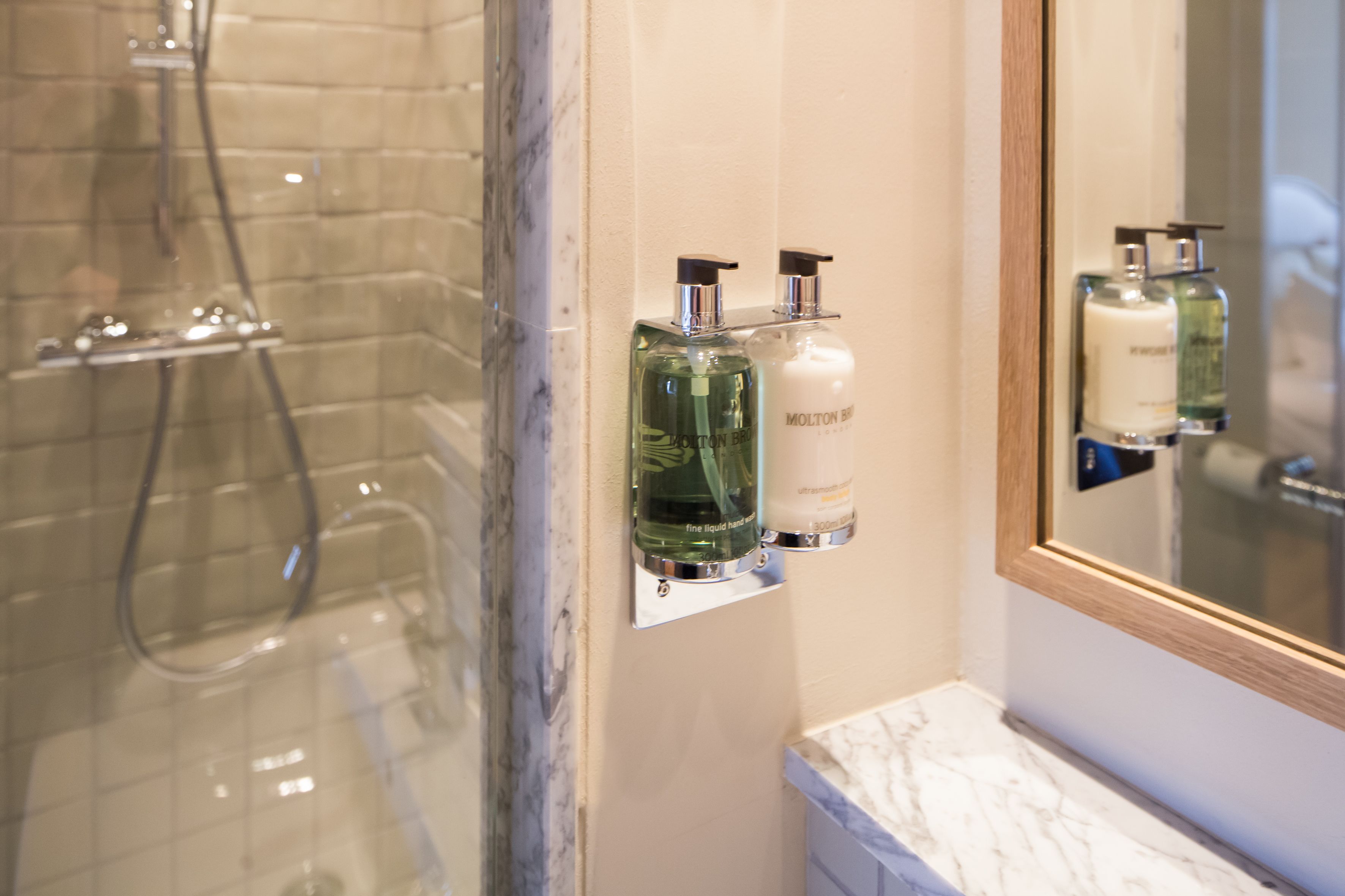 Un baño con mampara de vidrio, encimera de mármol y dos botellas de loción en un estante fijado a la pared.