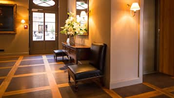A hotel lobby with a checkered floor, a reception desk, a chair, a vase with flowers, and a mirror on the wall.