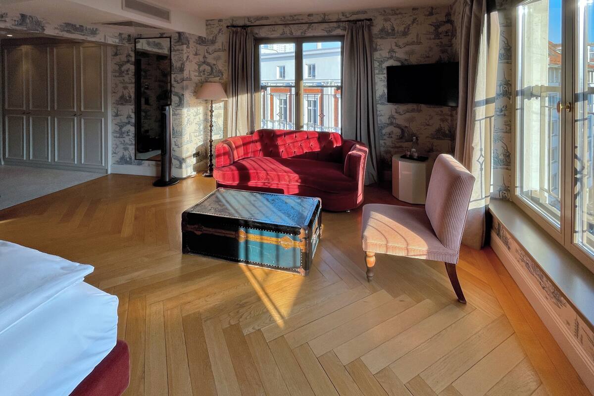 A spacious room with a red sofa, a wooden floor, a vintage suitcase, and a view of buildings through large windows.