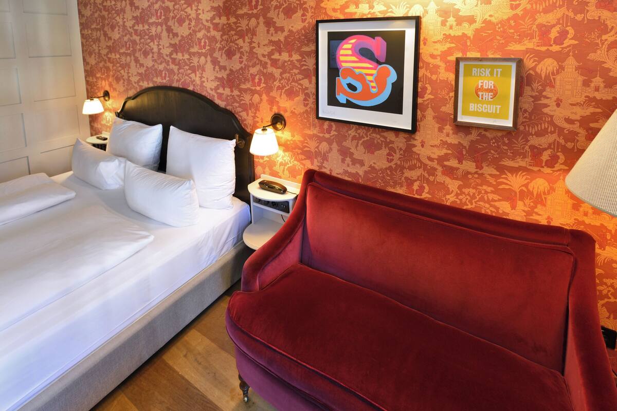 A hotel room with a bed, a red sofa, a nightstand with a lamp, and a wall with floral wallpaper and two framed pictures.