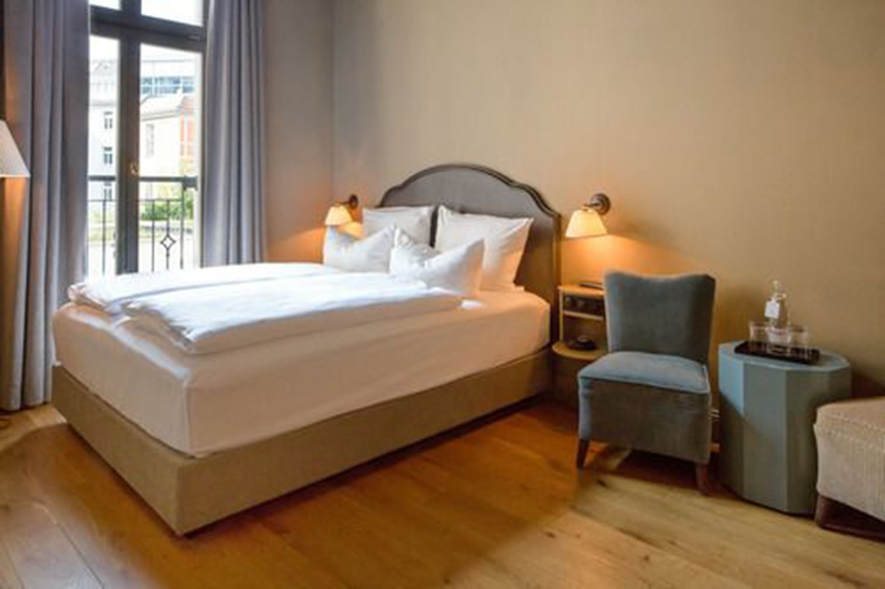Una cama bien tendida con sábanas blancas, una silla y una mesita de noche con una lámpara.