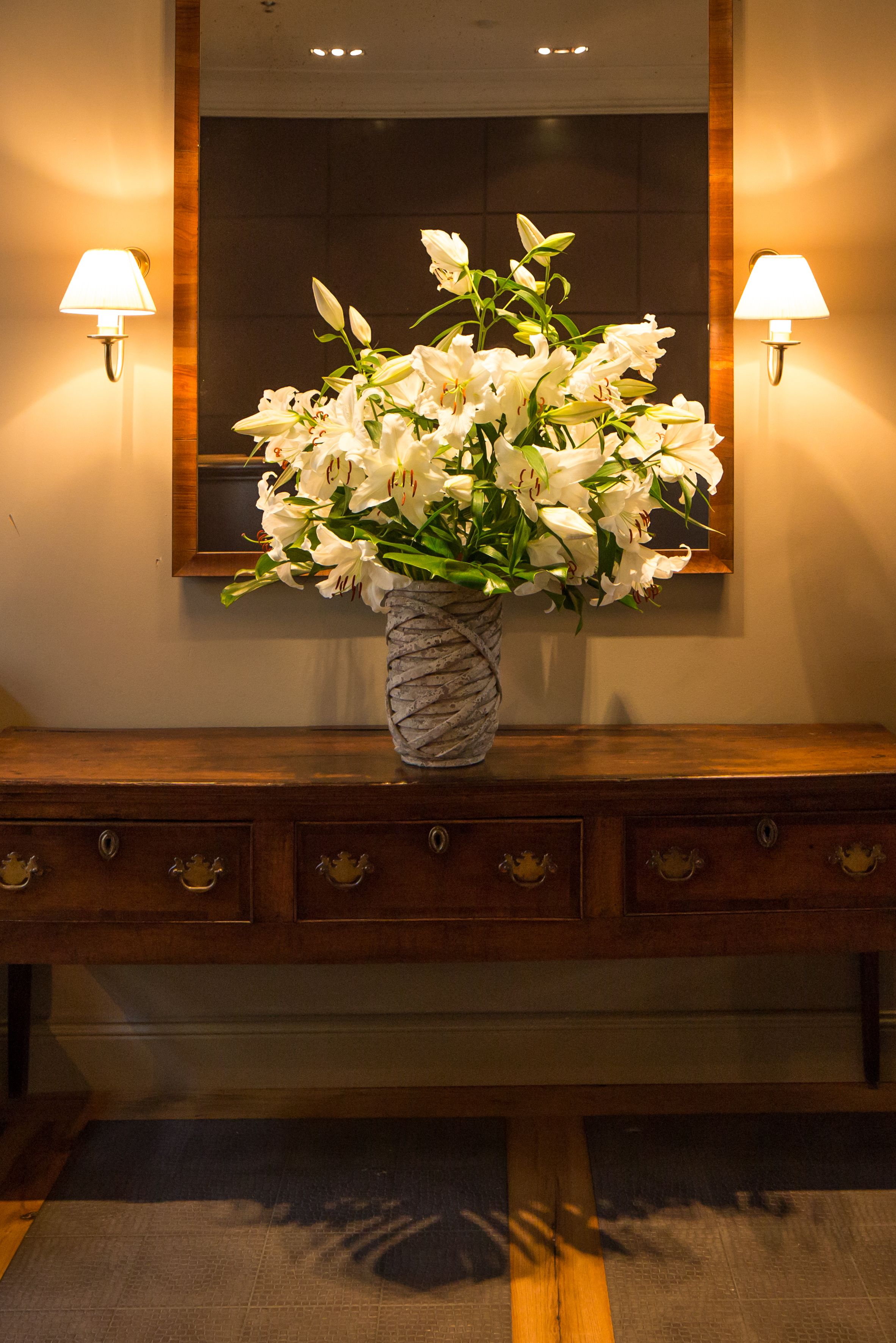 Un jarrón de mármol con flores blancas sobre una consola de madera con dos lámparas de pared.