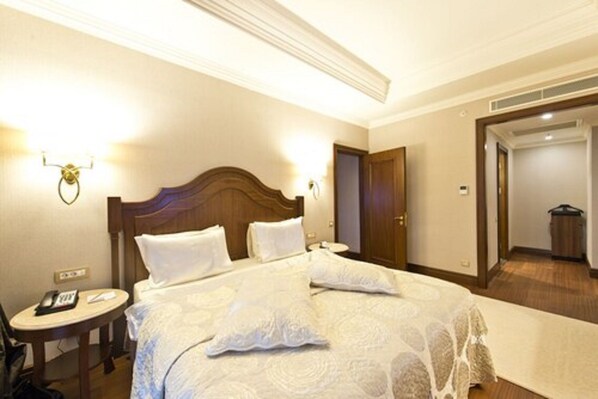 Una habitación de hotel con una cama grande, una mesita de noche con lámpara y una puerta que da a otra habitación.