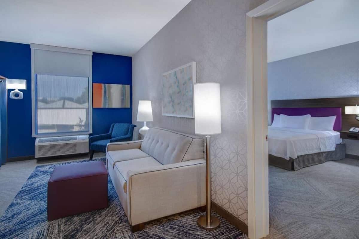 Habitación de hotel con sofá, mesita de noche, lámpara, cama y ventana con persianas.