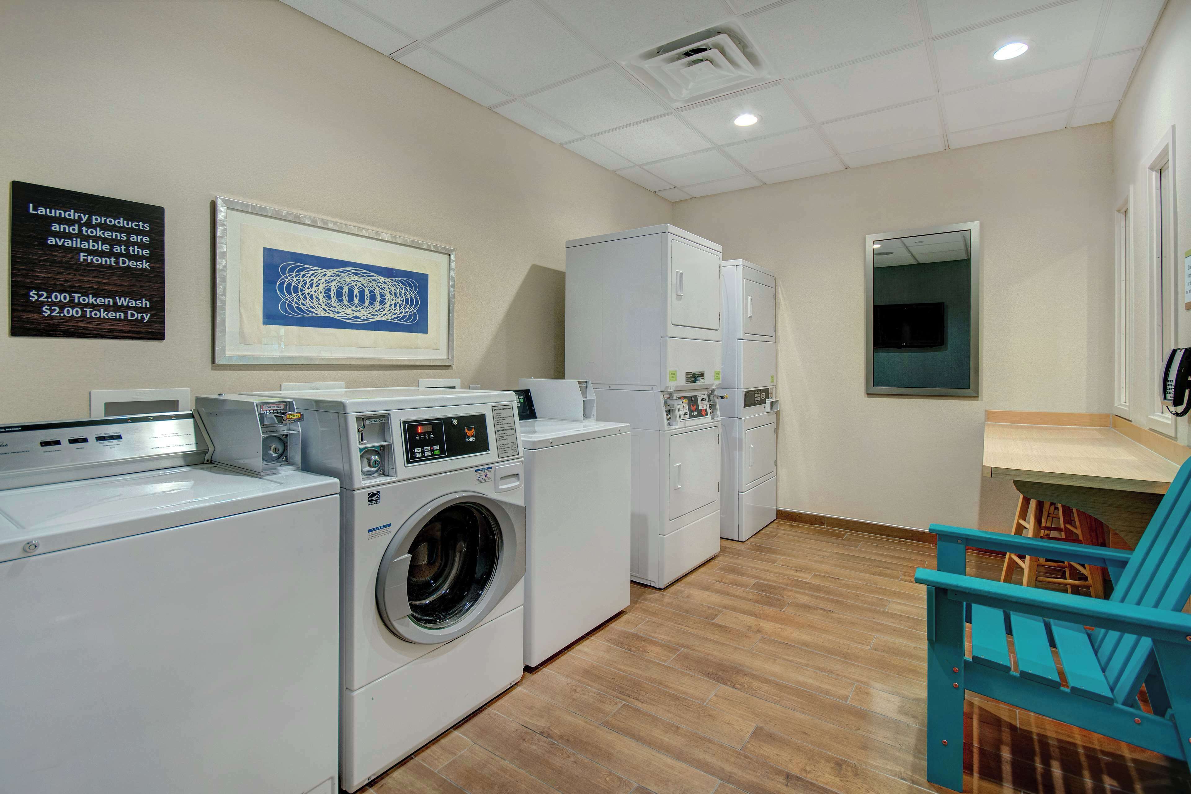 A laundry facility with washers and dryers, a sign for laundry products and tokens, and a framed artwork on the wall.