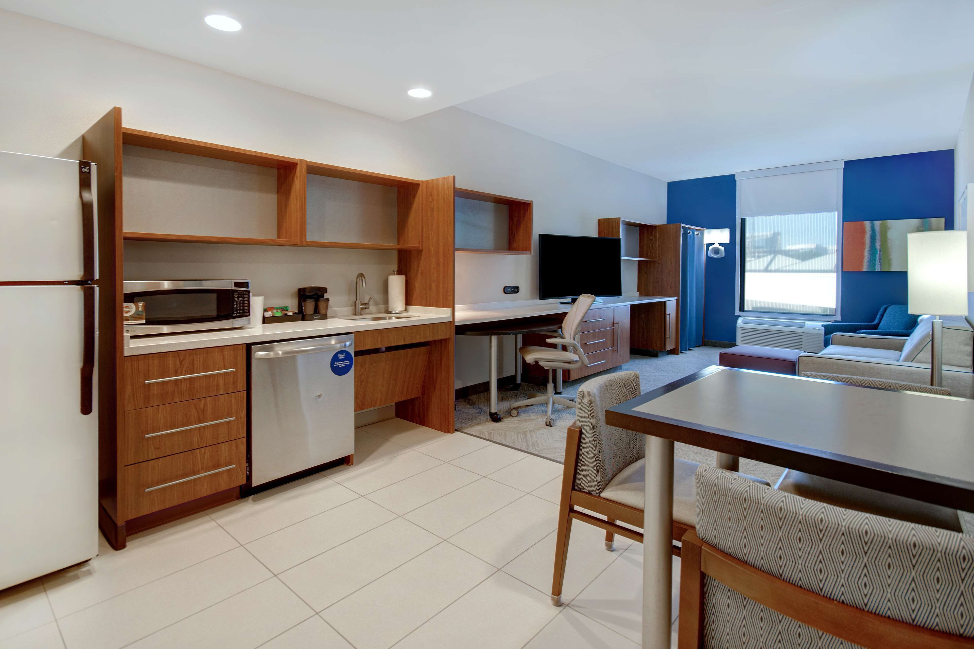 Una cocina moderna con microondas, refrigerador y armarios de madera. Hay una mesa de comedor con sillas y un televisor al fondo.