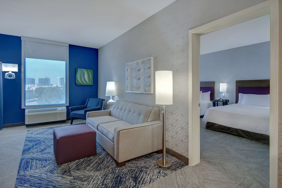 Habitación de hotel con sofá, cama, lámpara de pie y un amplio ventanal con persianas.