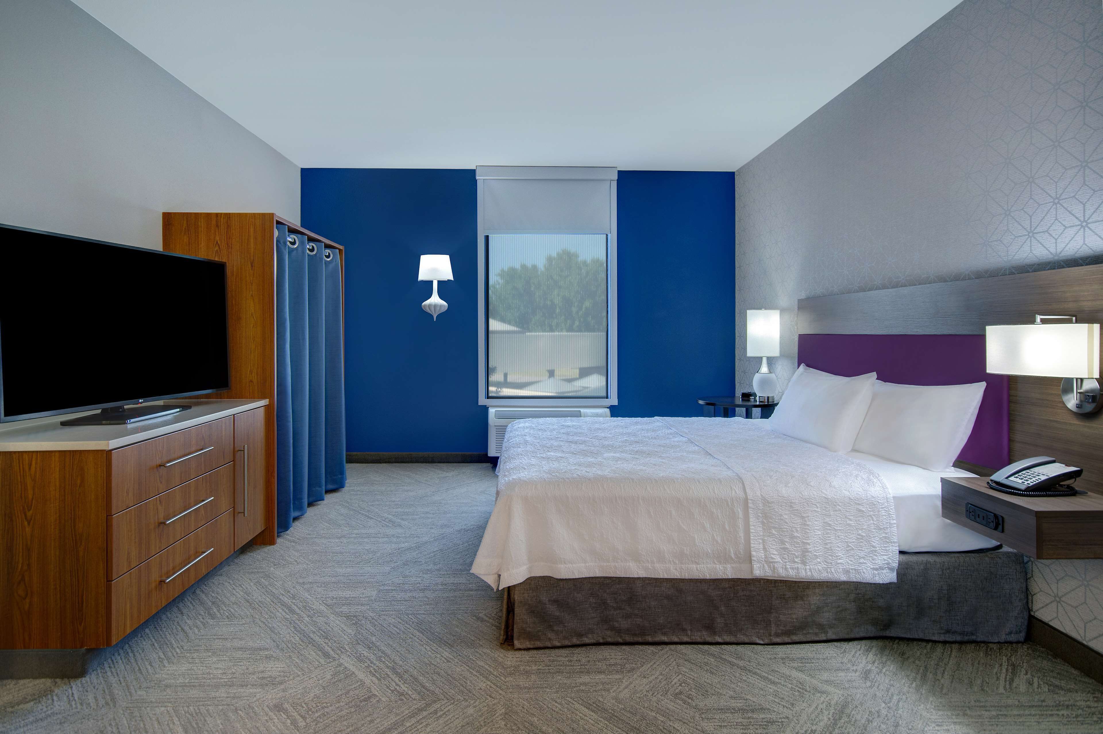 Une chambre d’hôtel dotée d’un grand téléviseur à écran plat, d’un lit avec du linge de lit blanc, d’une table de chevet avec une lampe et d’une fenêtre avec des rideaux bleus.