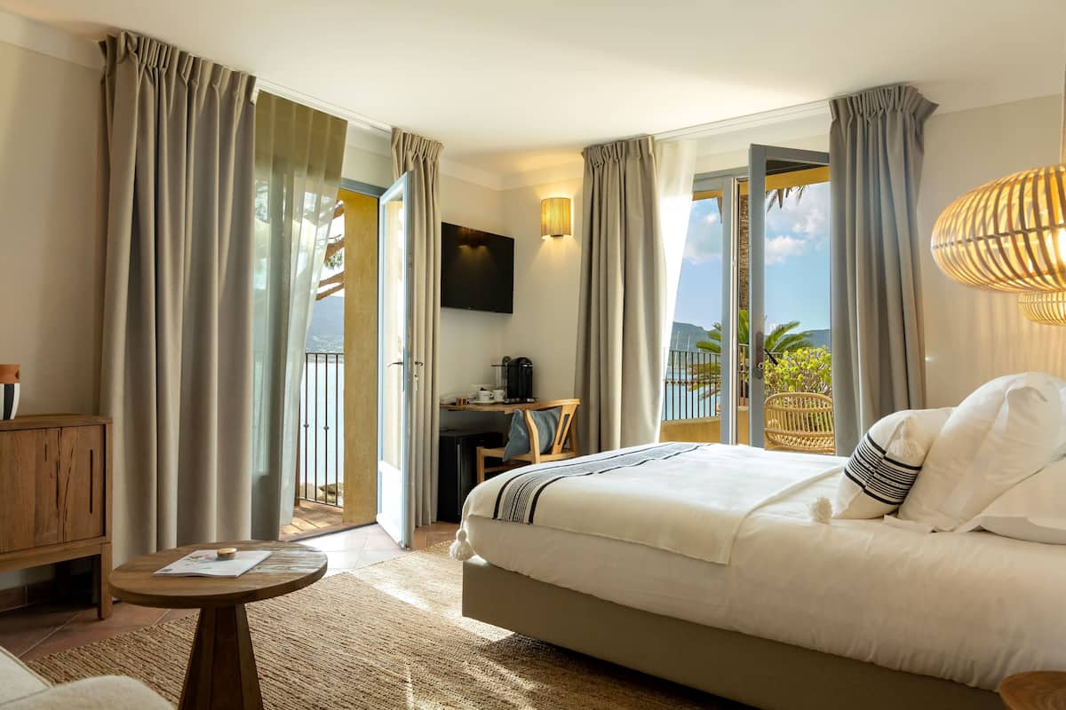 A hotel room with a large bed, a desk with a chair, a television, and a balcony with curtains.