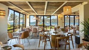 A restaurant with large windows offering a view of the sea and hills. Tables are set with chairs and dining ware. - Casa Santini x Roc Seven (Porto-Vecchio)