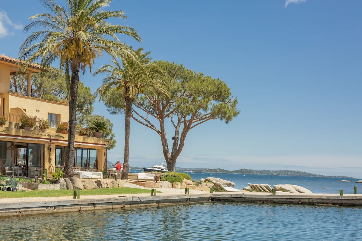 A waterfront property with a pool, palm trees, and a building with outdoor seating.