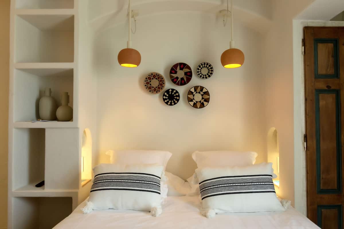 A neatly made bed with white bedding and striped pillows, a shelf with decorative items, and a wall adorned with patterned plates.