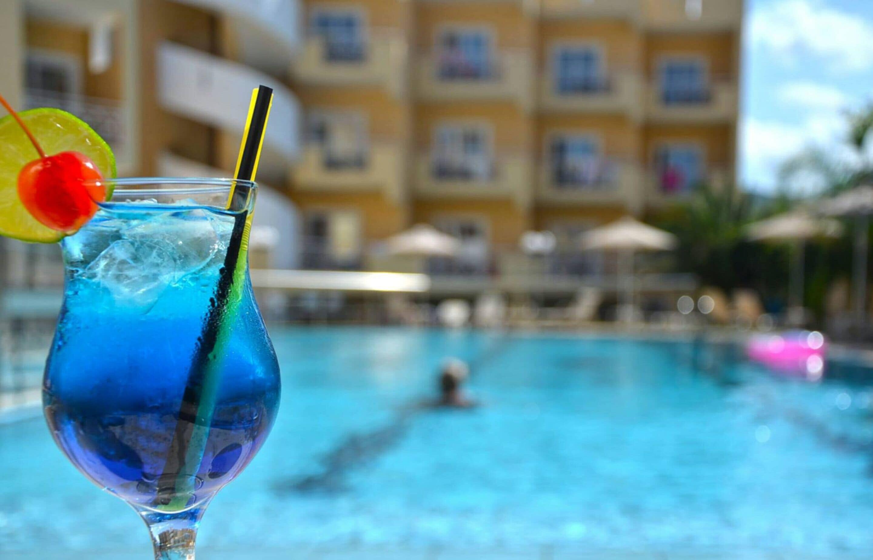 Un verre contenant un liquide bleu, une paille et une cerise posée sur une tranche de citron vert, placé devant une piscine où une personne se trouve dans l’eau, avec un bâtiment d’hôtel en arrière-plan.