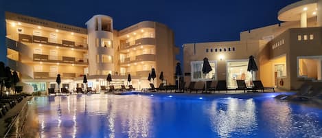 Una piscina con sillas de descanso y sombrillas, rodeada de edificios de varios pisos con balcones y ventanas iluminadas.