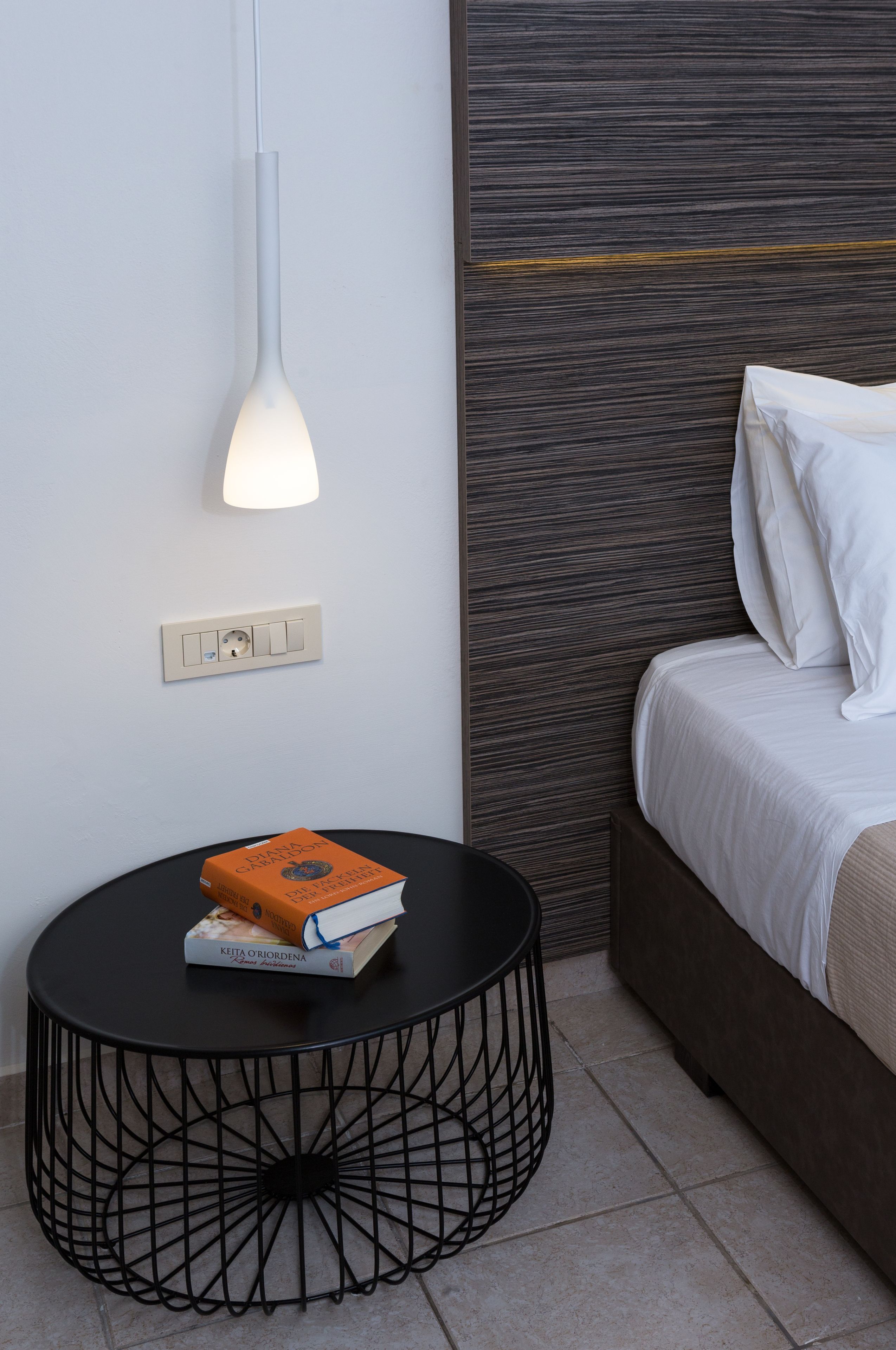 A modern bedroom with a bed, a round black table with a book, a wall-mounted light, and a power outlet.