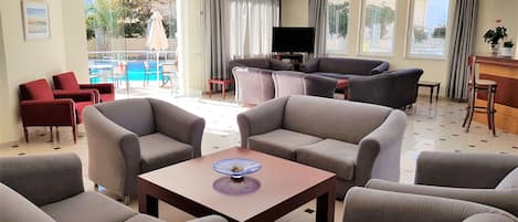 A modern living room with grey sofas, a coffee table, and a TV. There is a pool visible through the glass doors.