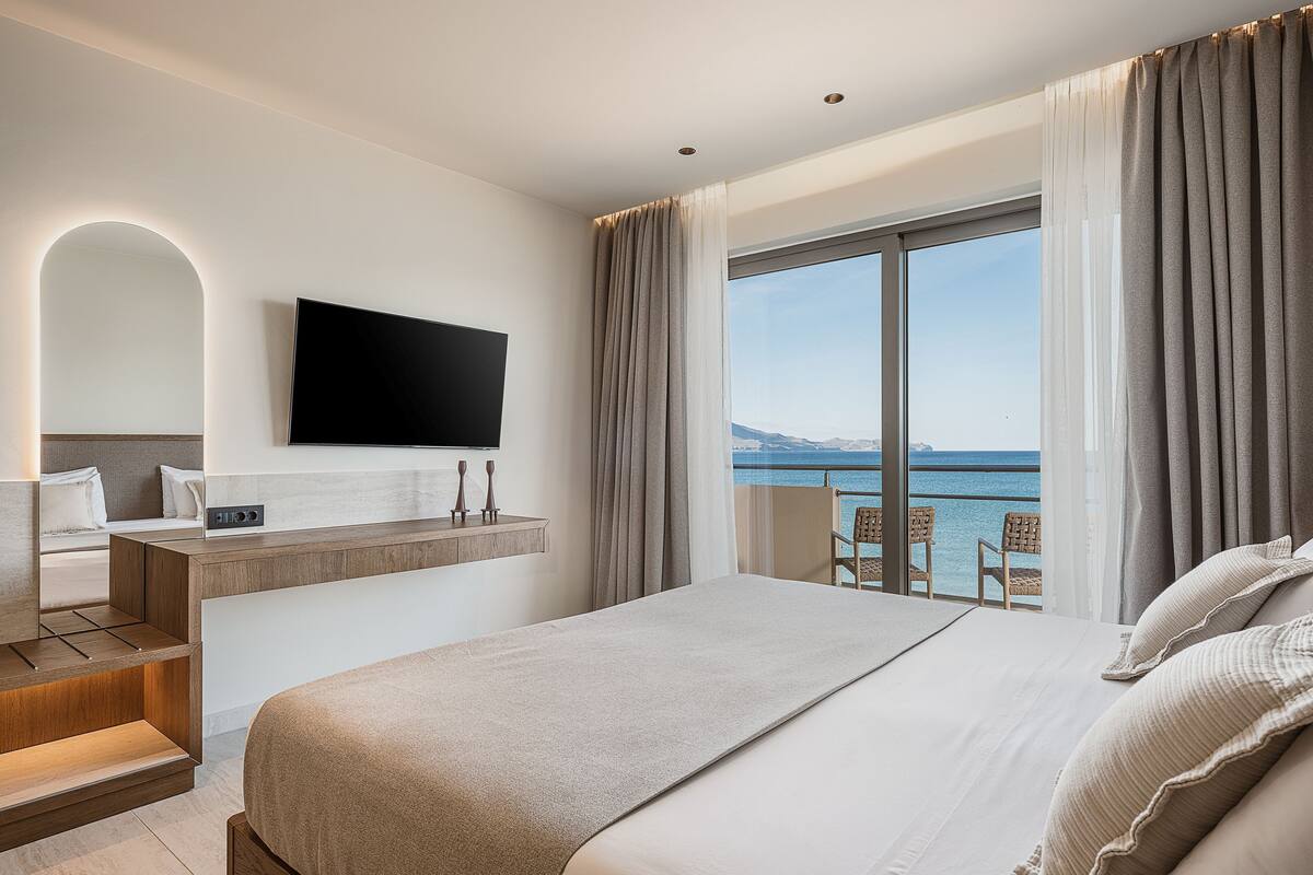 Une chambre d’hôtel moderne dotée d’un grand lit, d’un téléviseur fixé au mur, d’un bureau avec une chaise et d’un balcon offrant une vue sur la mer.