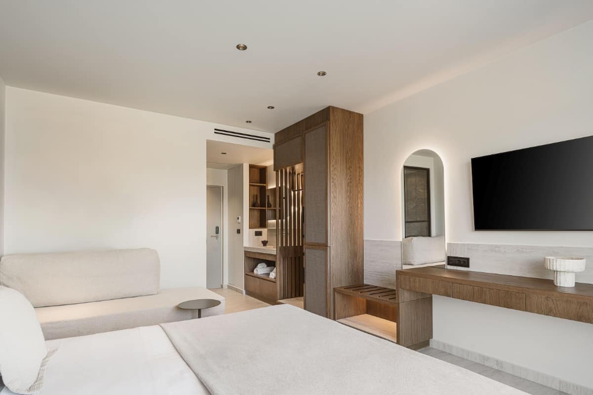Une chambre d’hôtel moderne comprenant un lit, un canapé, une petite table ronde, une armoire en bois, un téléviseur intégré et une salle de bain avec douche et lavabo.