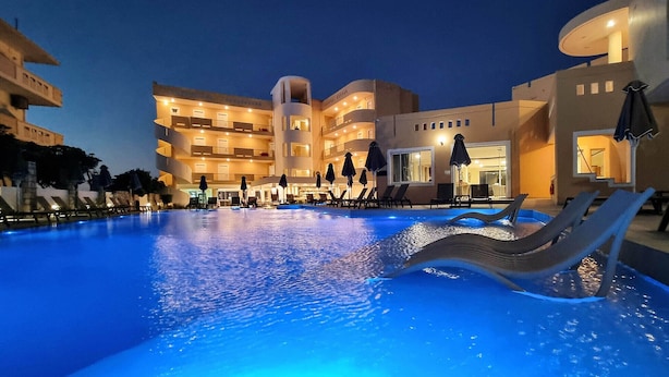 Una piscina con sillas de descanso y sombrillas, rodeada por un edificio de varios pisos con balcones y un bar junto a la piscina.
