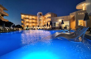 A swimming pool with lounge chairs and umbrellas, surrounded by a multi-story building with balconies and a poolside bar. - Sunny Bay Hotel (Kissamos)