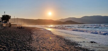 Una playa con costa rocosa, algunas personas sentadas en la arena y montañas al fondo.