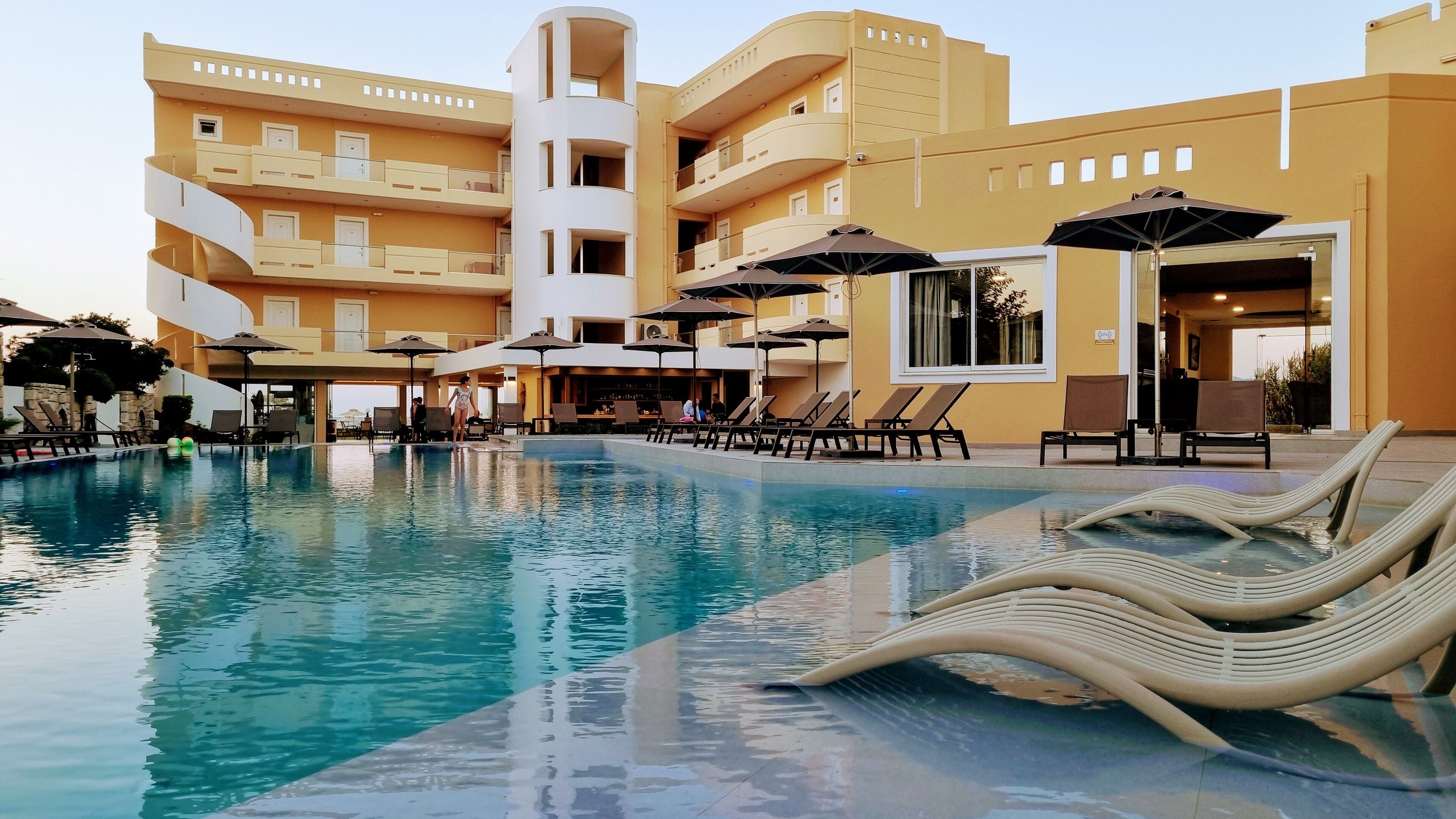 A hotel pool area with lounge chairs and umbrellas, a bar area, and a multi-story building with balconies.