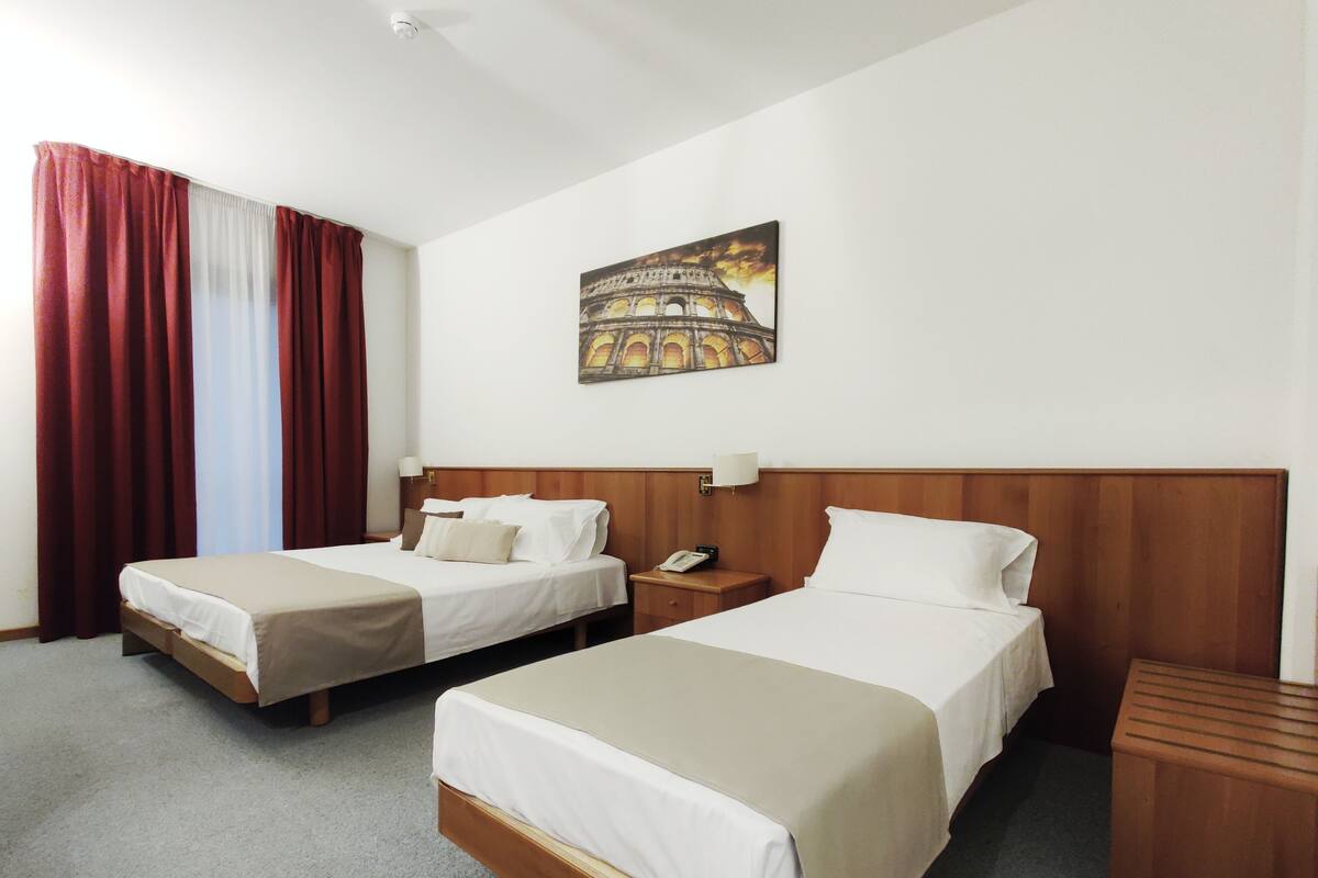 A hotel room with two beds, a nightstand with a phone, a painting of the Colosseum, and a window with red curtains.