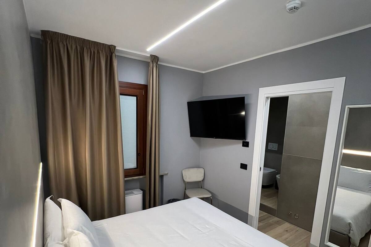 A hotel room with a bed, a TV mounted on the wall, a chair, a toilet, and a mirror.