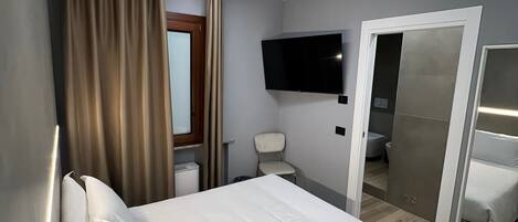 Habitación de hotel con cama, televisor en la pared, una silla, inodoro y espejo.