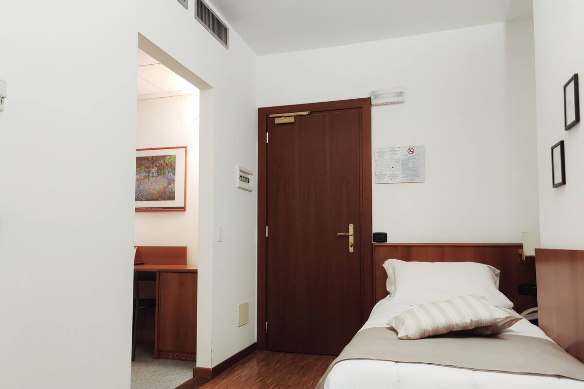 A hotel room with a wooden door, a bed with white bedding, a desk with a chair, and a framed picture on the wall.